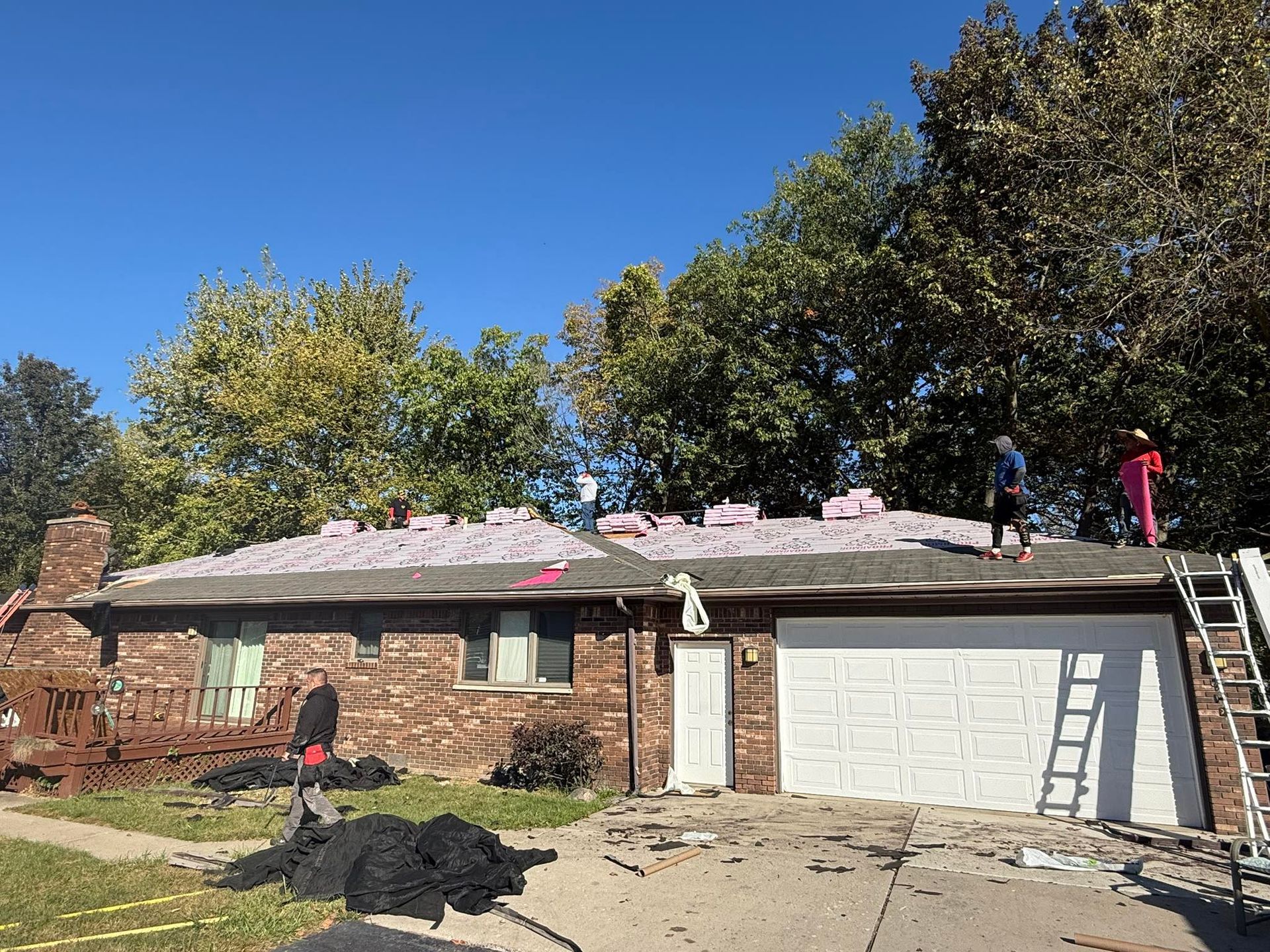Roofing workers on a house, tearing off old shingles. Roof partially bare, with new materials in place. Blue sky.
