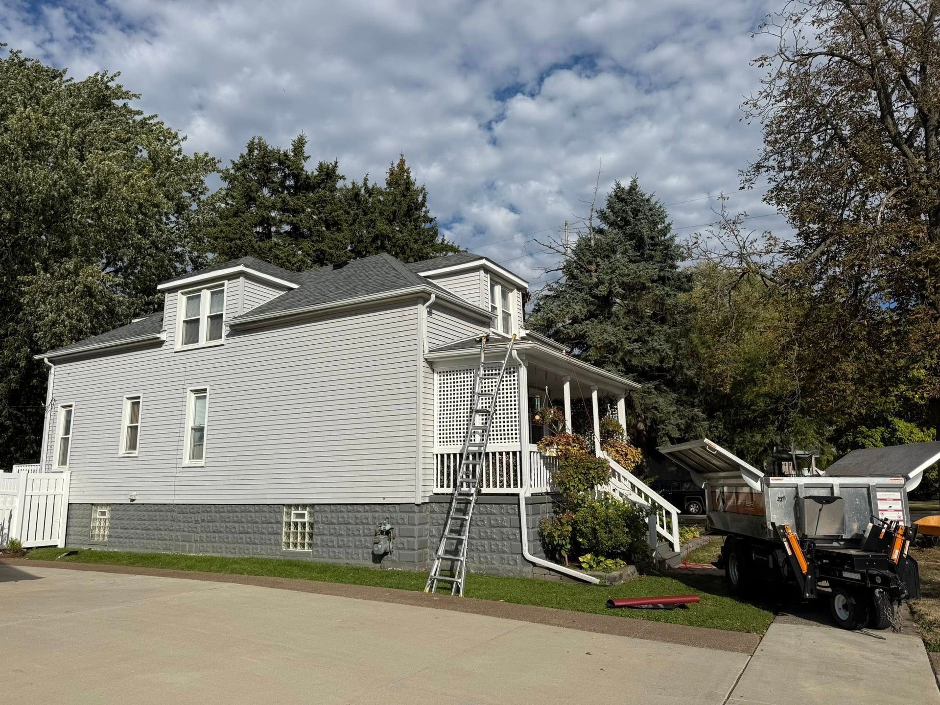 Gray house with white trim, porch, and a ladder against it; trees and sky in the background.