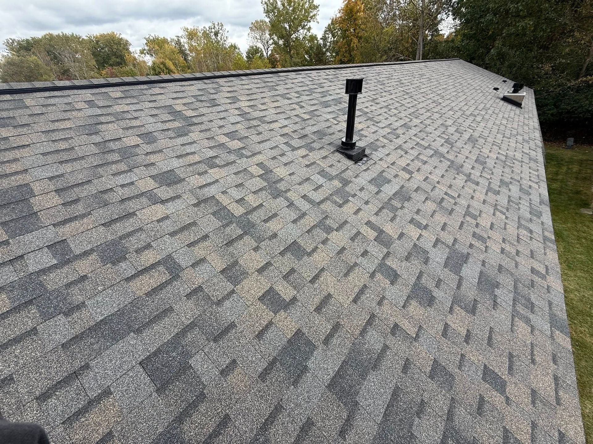Close-up of a shingle roof with black and gray asphalt shingles. A black vent pipe is visible.