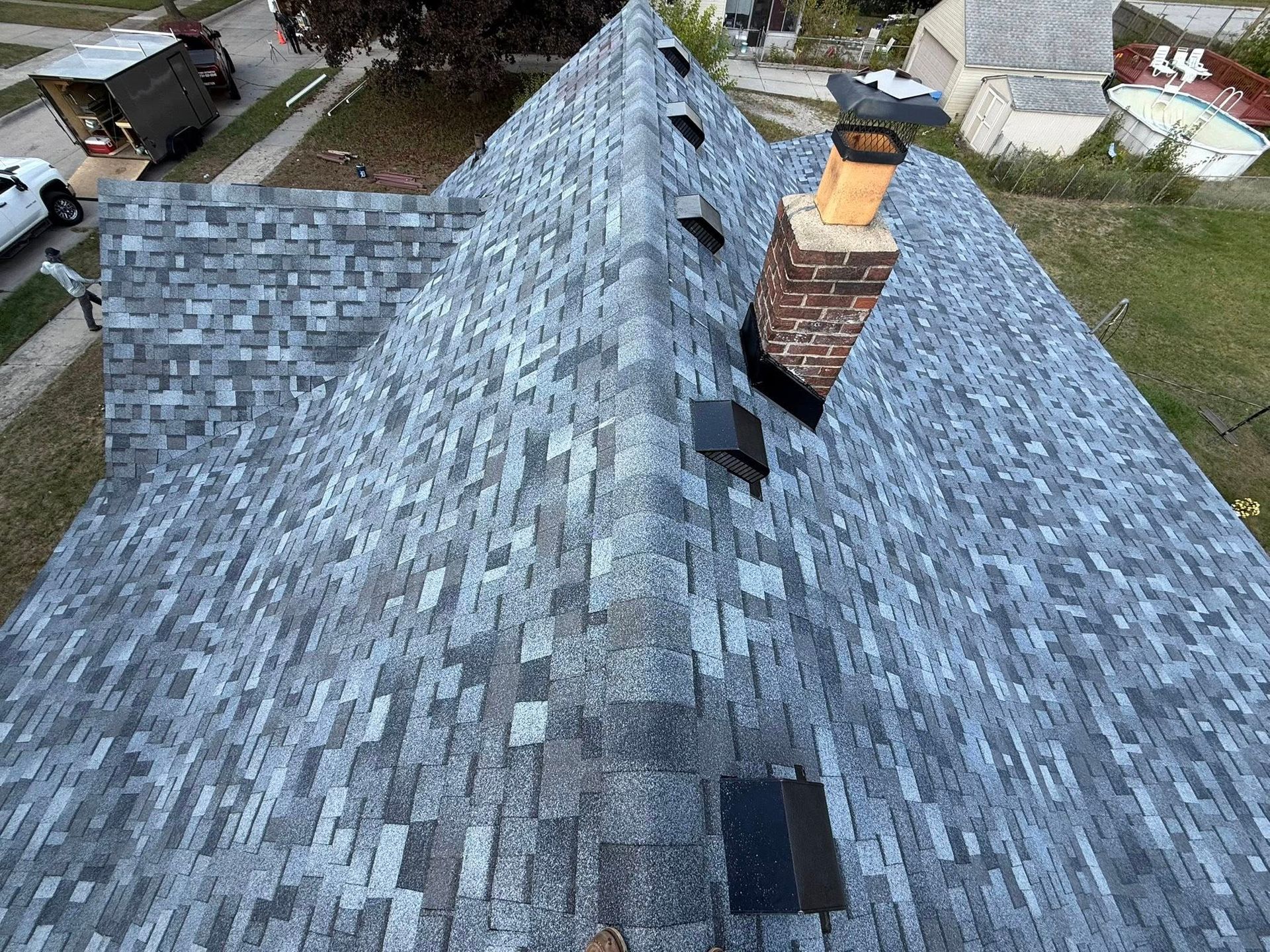 Overhead view of a house roof with grey shingles, chimney, and vents. A work truck and buildings are in the background.