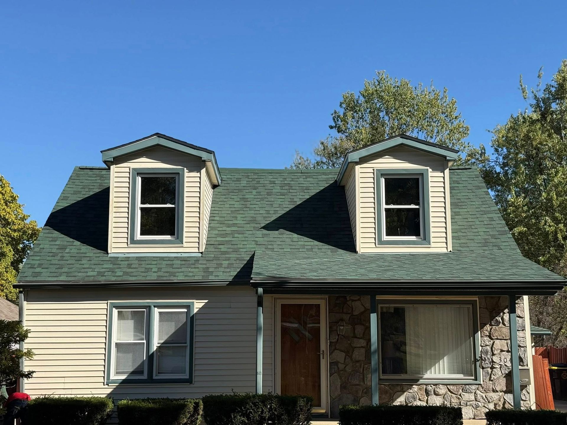 Tan house with green roof, two dormers, and stone accents, under a clear blue sky.