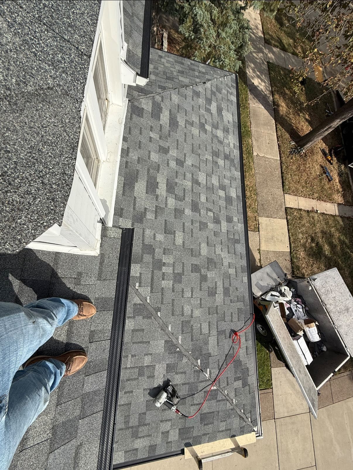 Person on a roof installing shingles; gray shingles and siding, tools nearby.