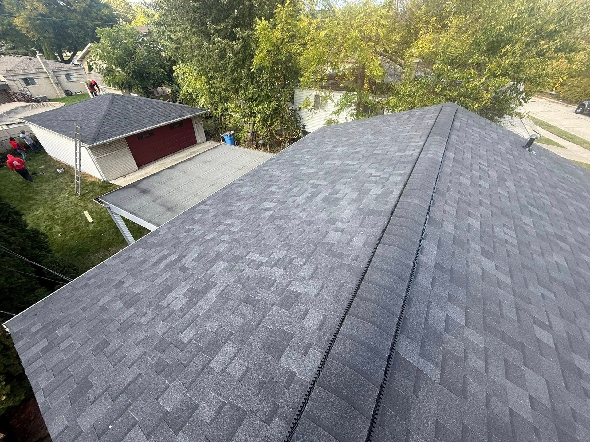 View from a roof of a house, looking at the dark gray shingles and surrounding green trees and other buildings.