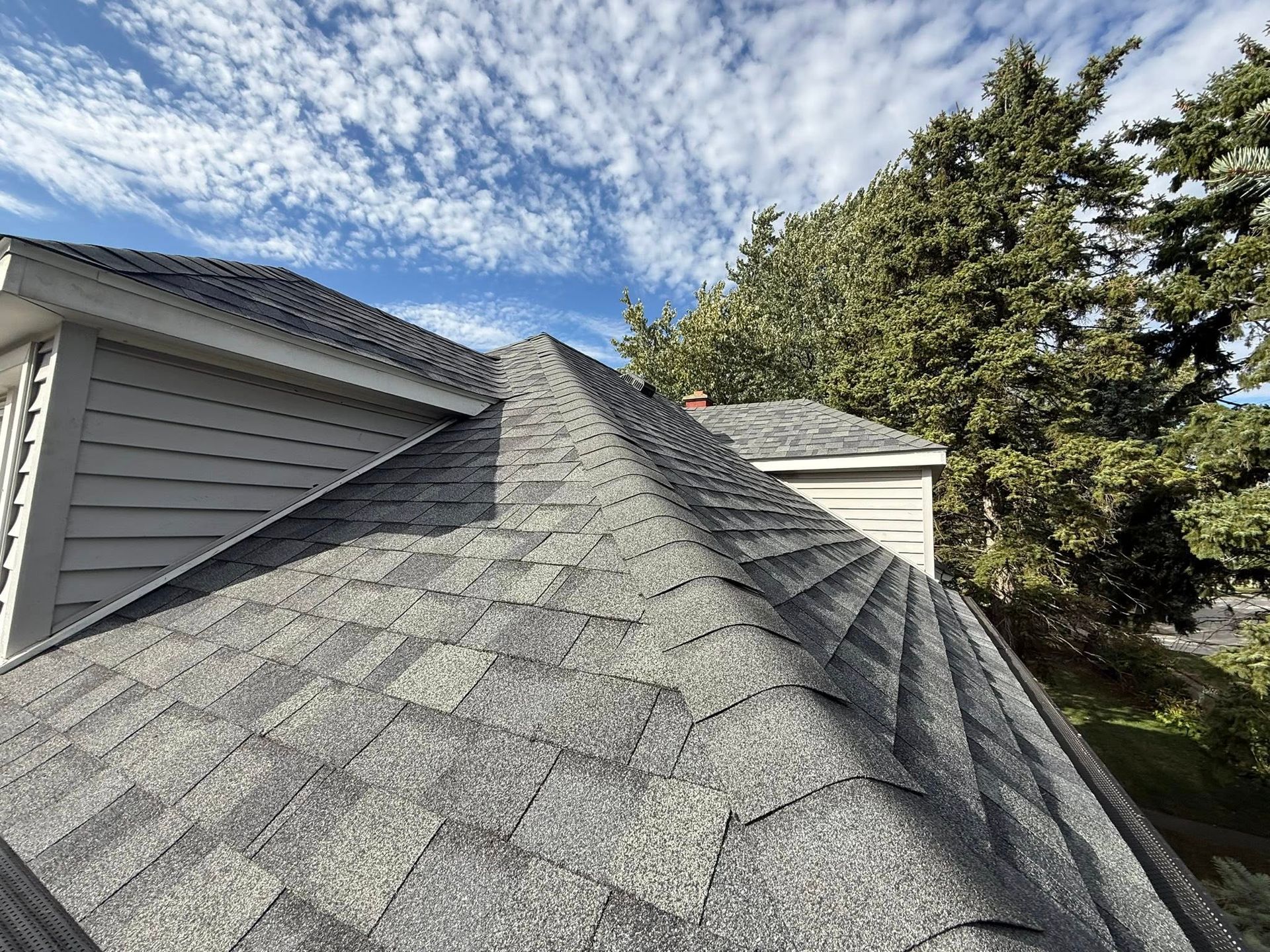 Gray asphalt shingle roof on a house, with a cloudy blue sky and trees in the background.