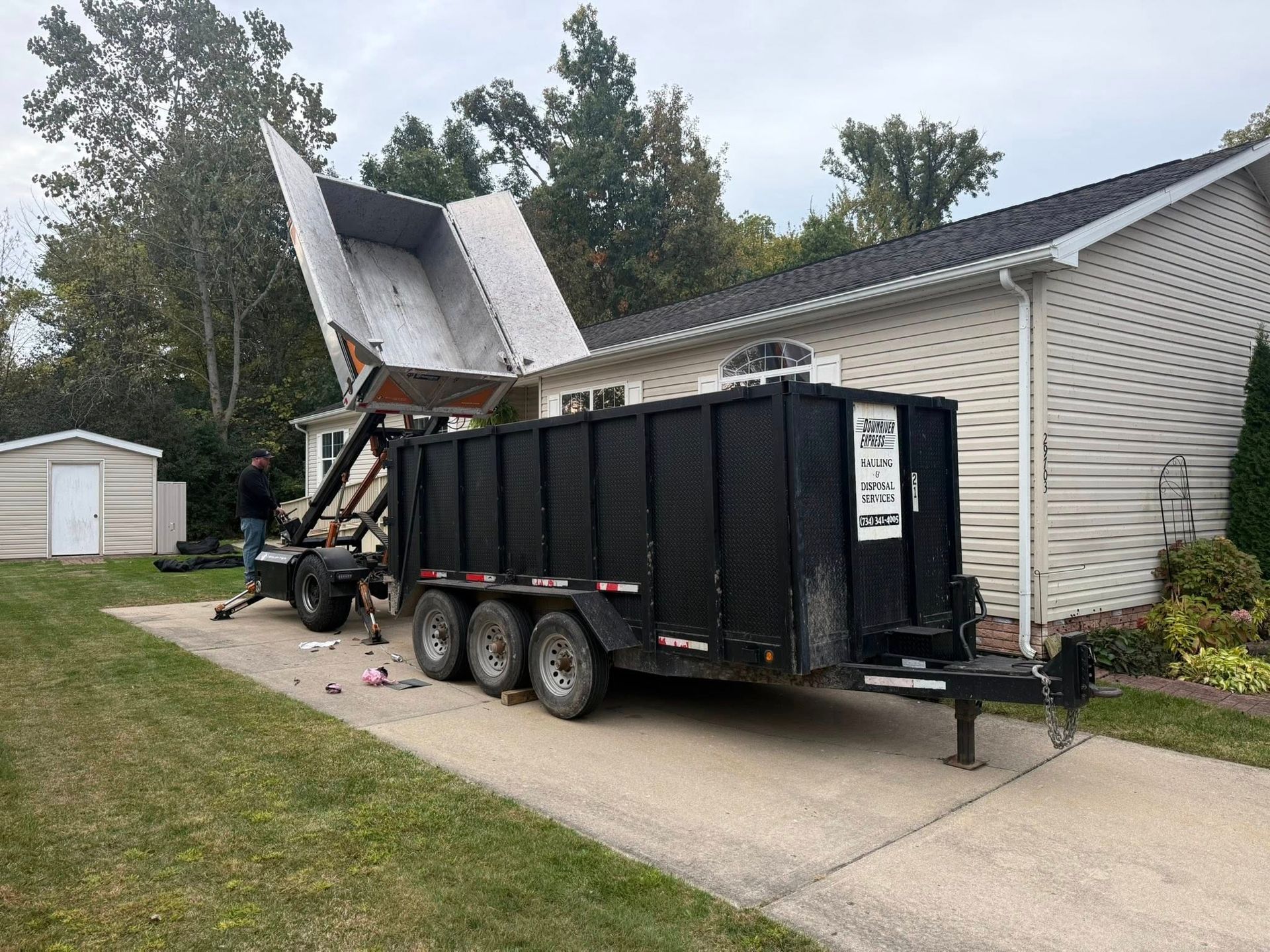 A black trailer with a tilted bed next to a house, dumping debris. A person stands nearby.