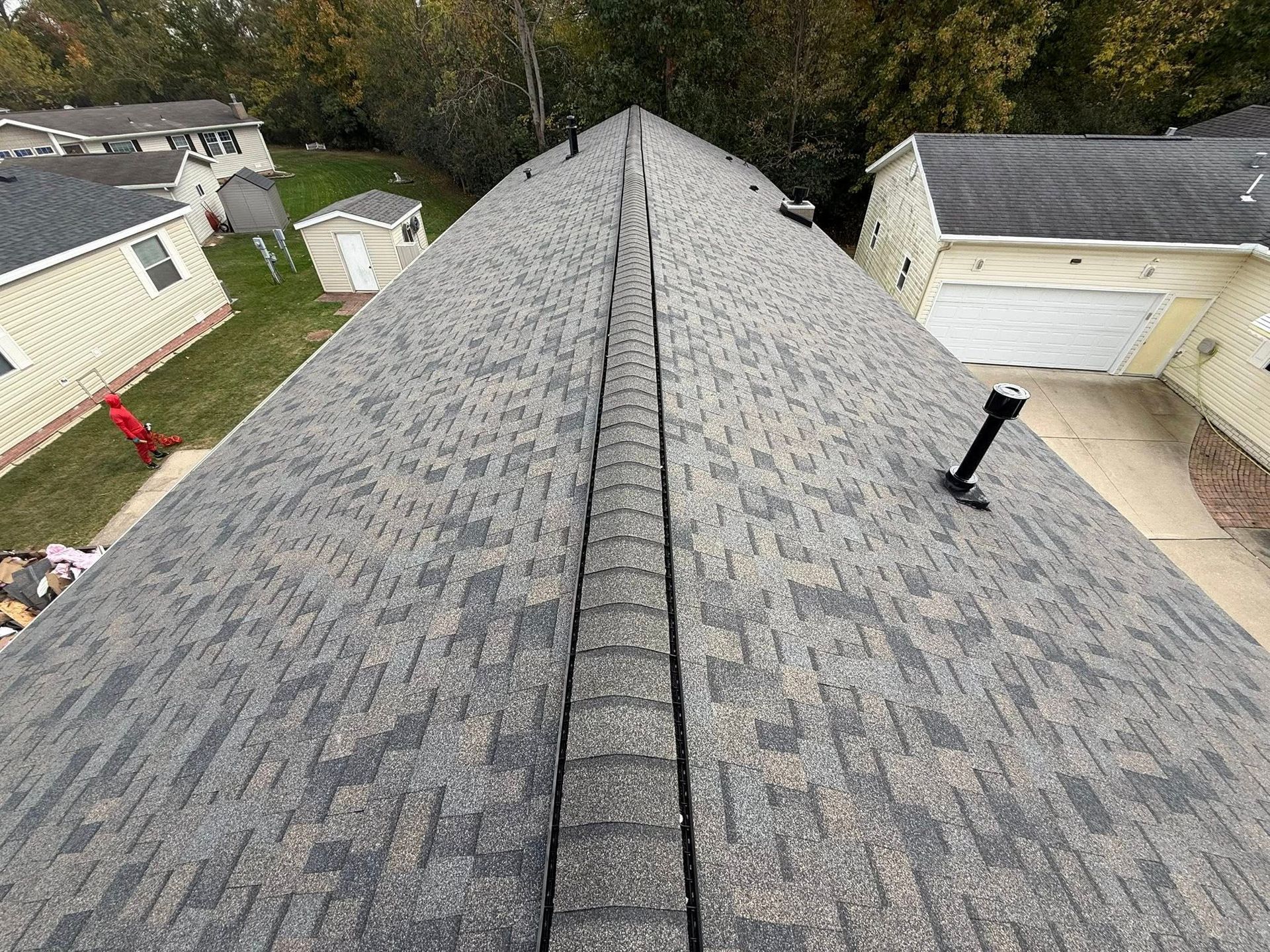 Overhead view of a gray shingled roof with a central ridge, surrounded by trees and houses.