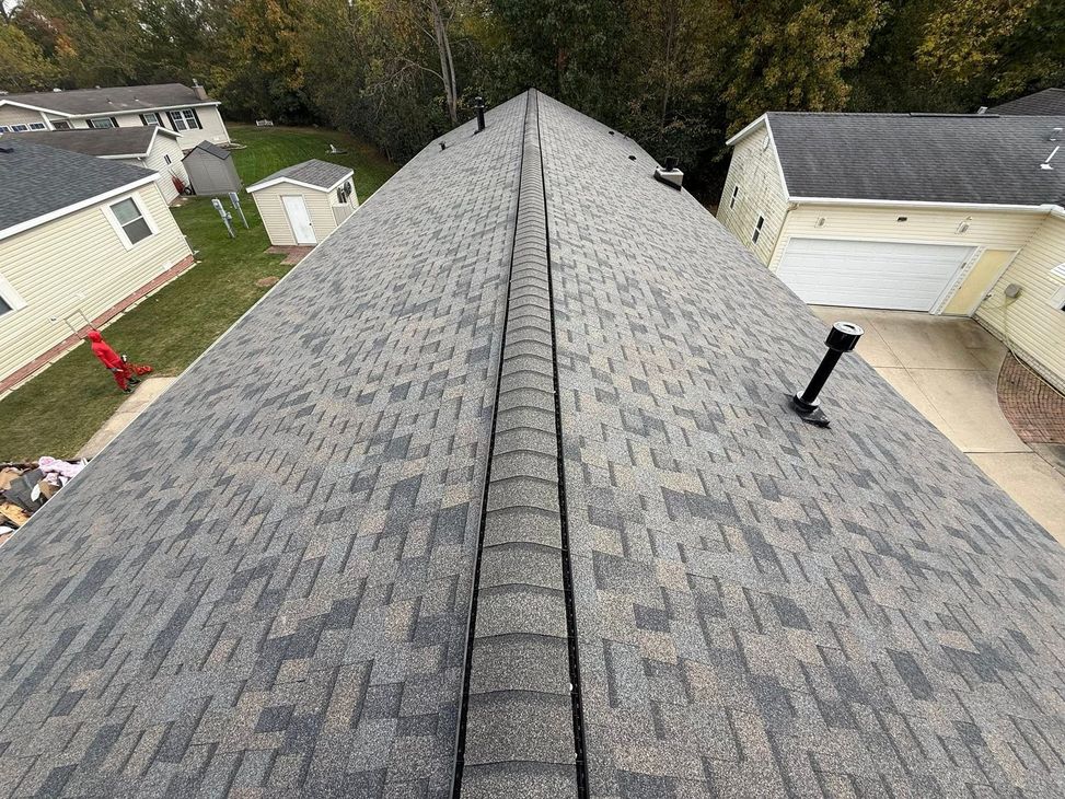 Overhead view of a gray shingled roof with a central ridge, surrounded by trees and houses.