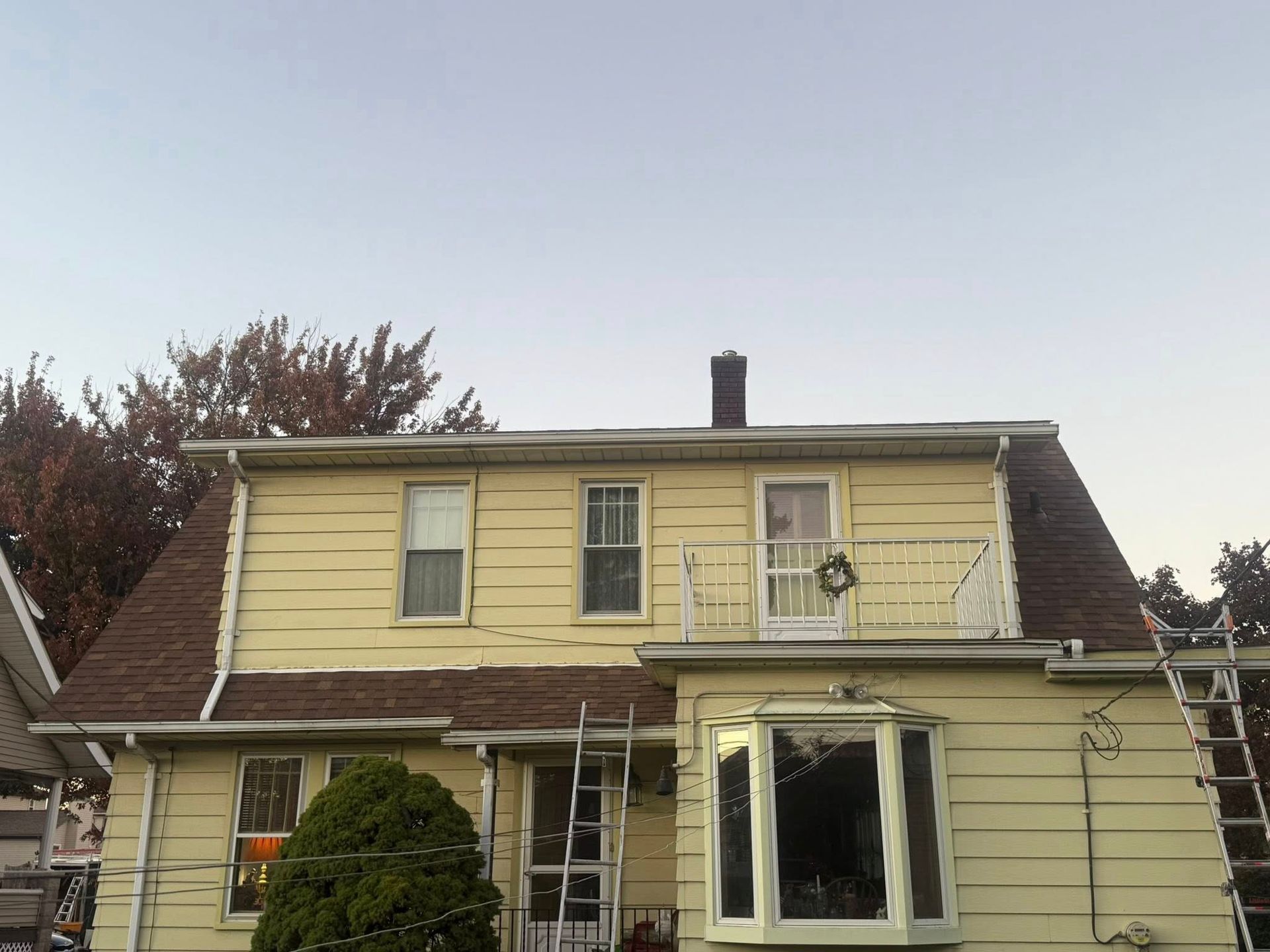 Two-story house with yellow siding, brown roof, a balcony, and ladders. Cloudy sky in the background.