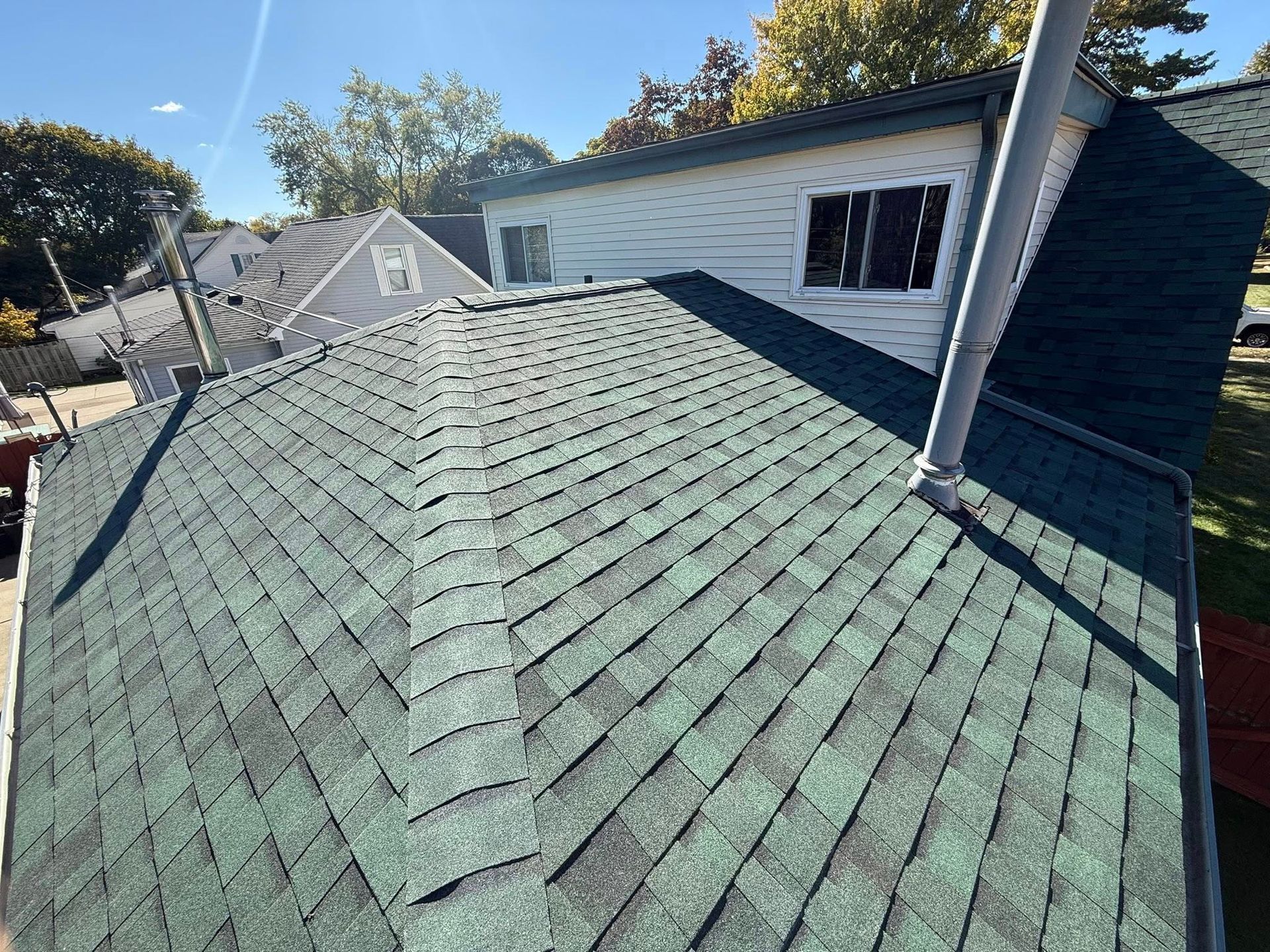 Green shingled roof with a chimney and surrounding houses on a sunny day.