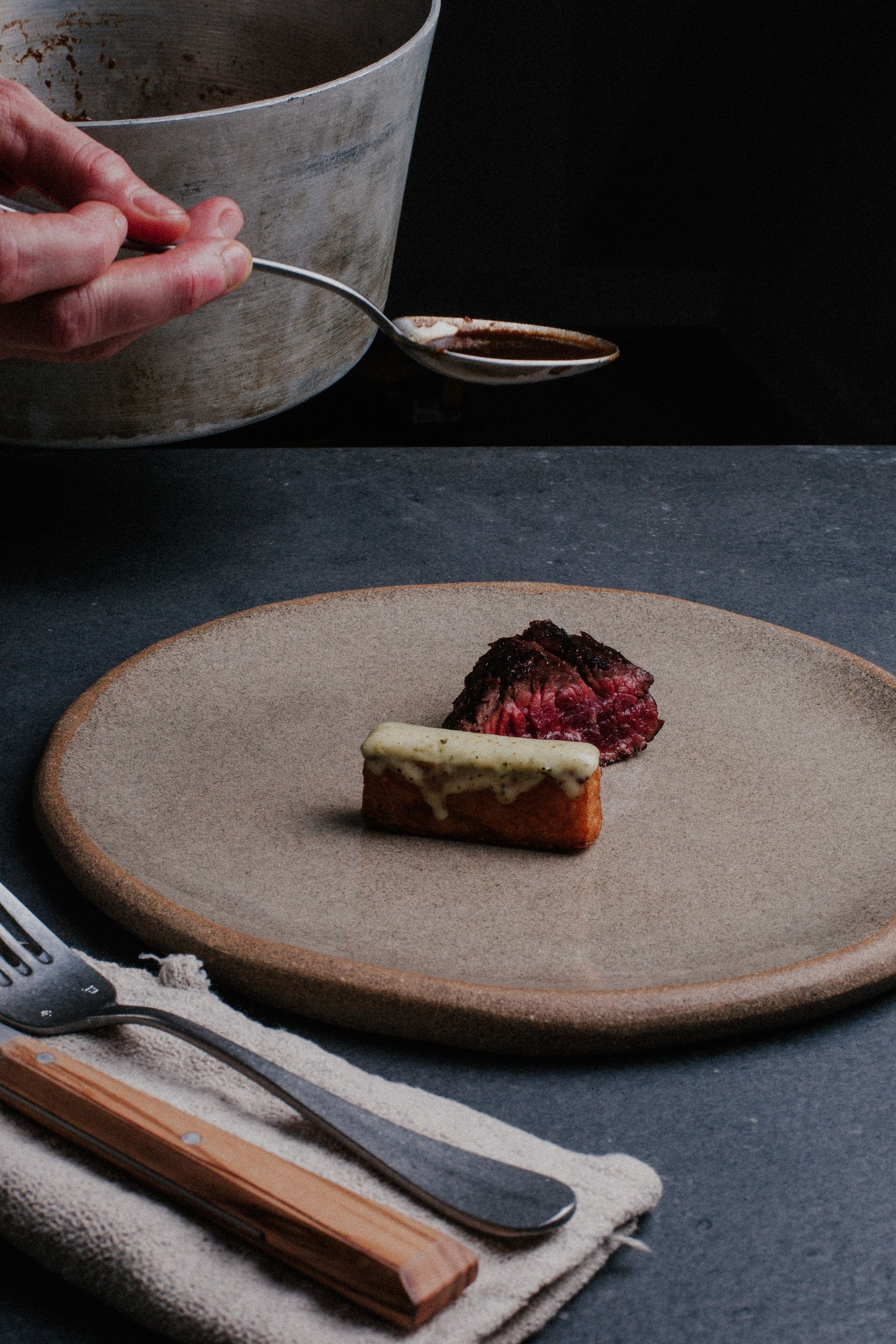 Plat gastronomique présenté dans une assiette, arrosé de sauce. Comprend une fourchette, une cuillère et un couteau à manche en bois.