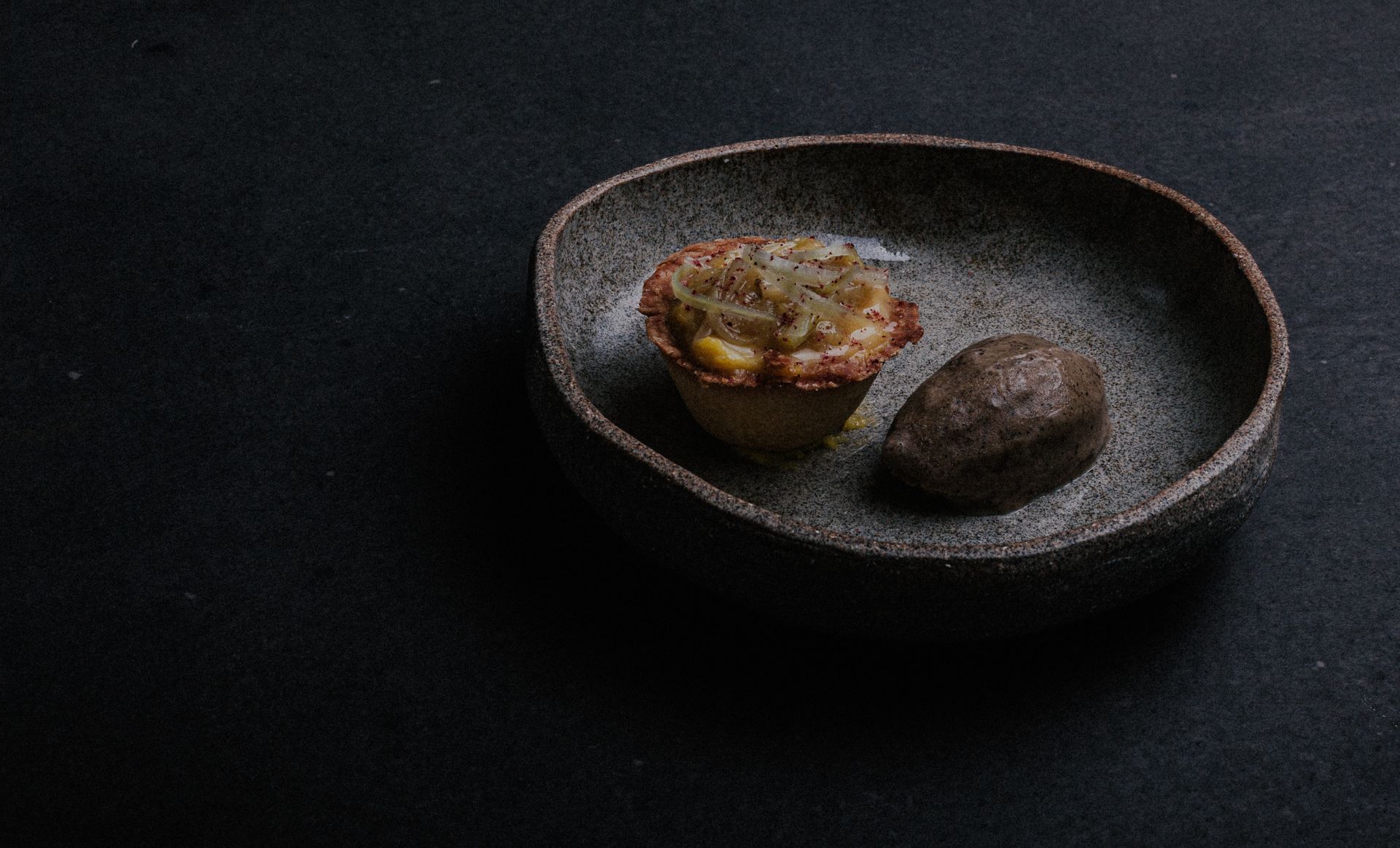 Une petite pâtisserie et un objet rond et sombre dans un bol rustique posé sur une surface noire.