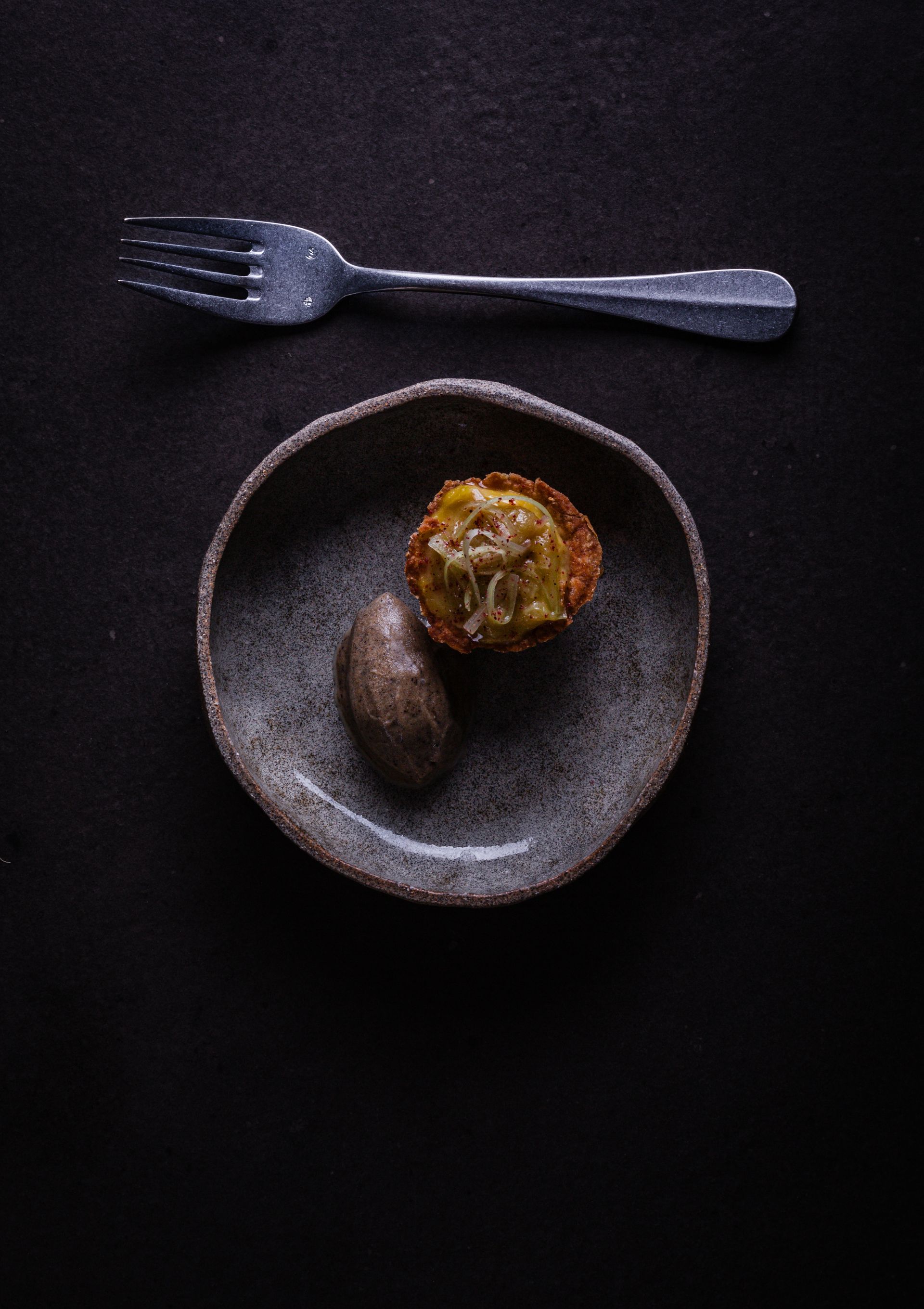 Petite tarte et aliment oblong sur une assiette grise avec une fourchette sur une surface sombre.