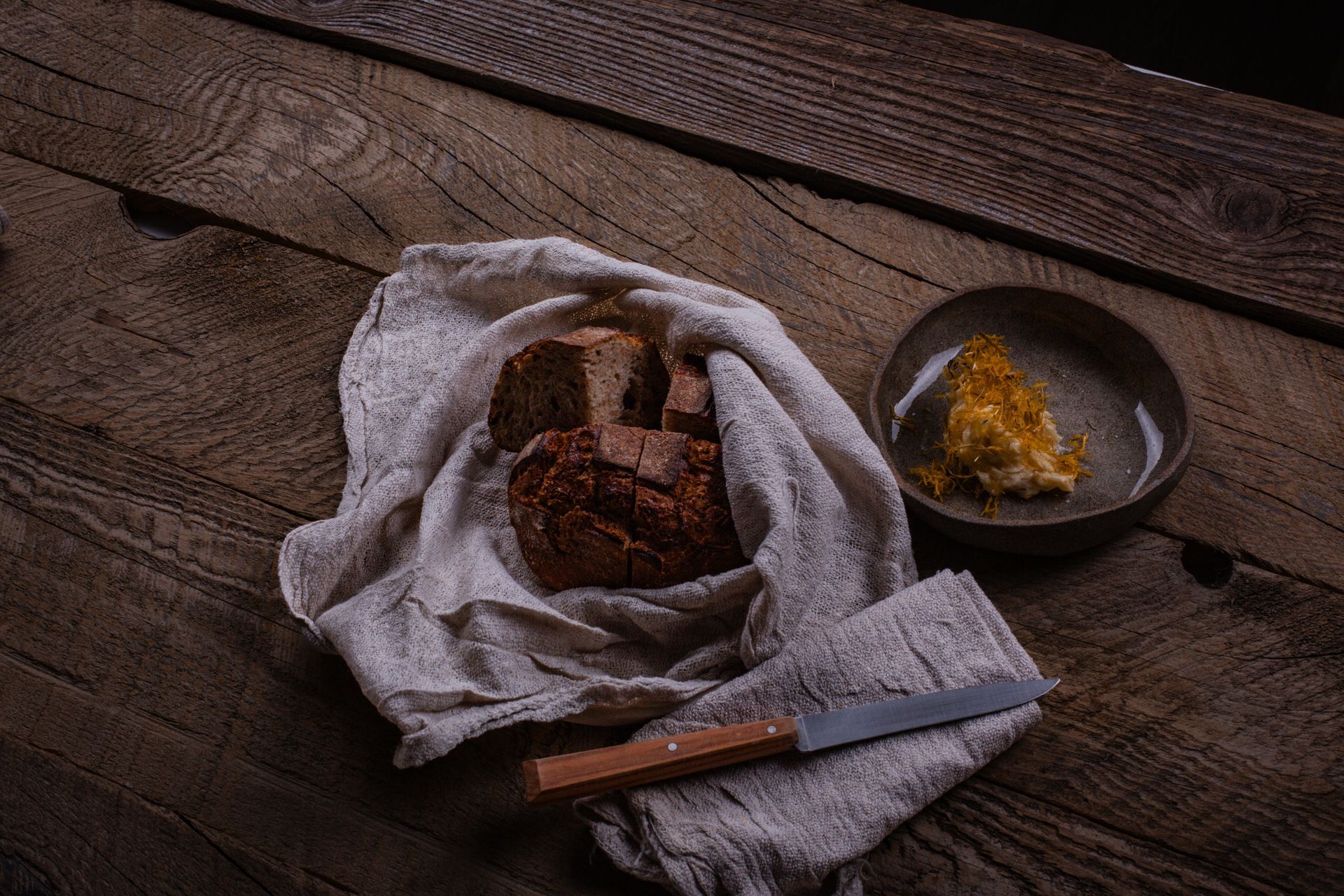 Du pain tranché enveloppé dans un torchon, du beurre dans un bol et un couteau sur une table en bois.