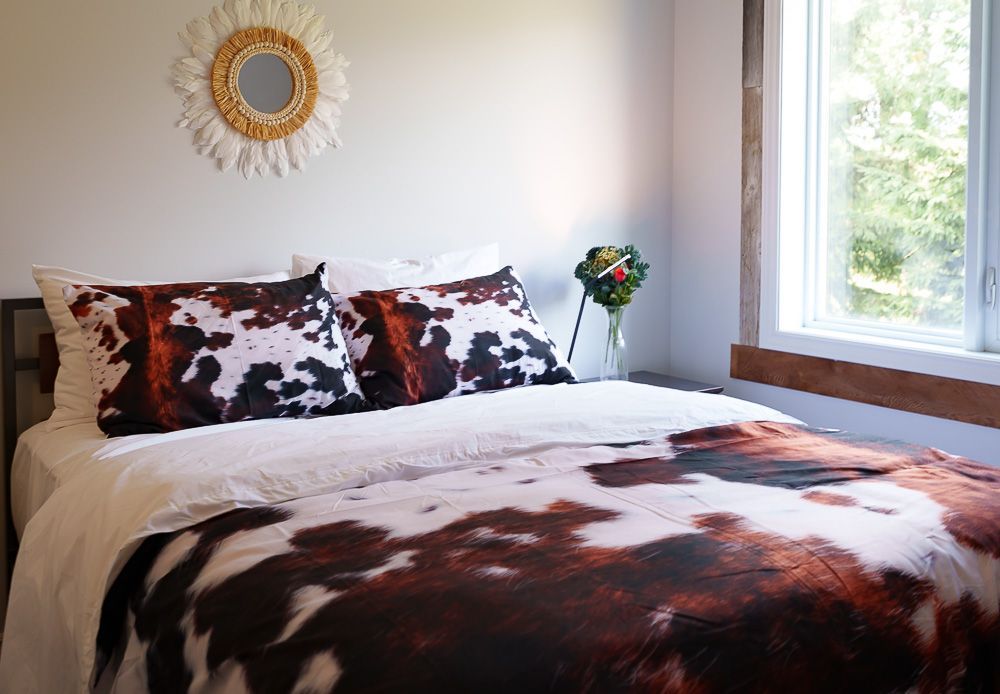 Chambre avec linge de lit à motifs peau de vache, murs blancs, miroir tissé et petit arrangement floral près de la fenêtre.
