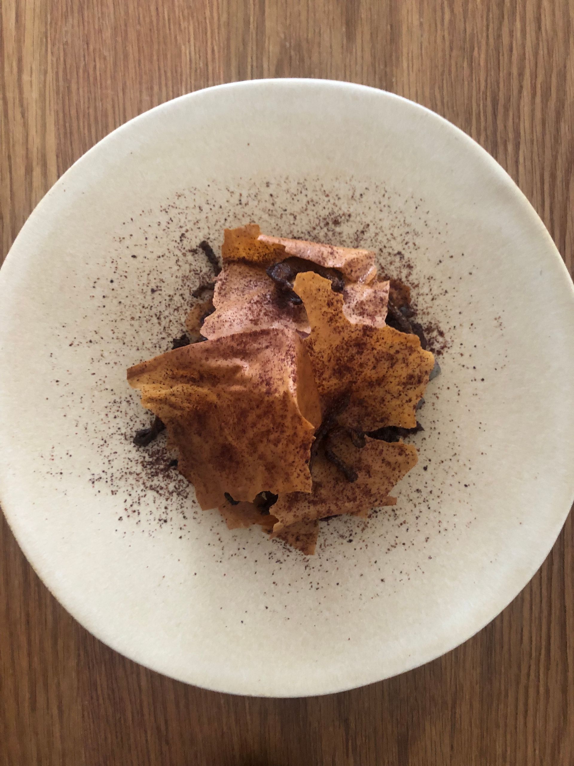 Dessert sur une assiette blanche : chips brun clair, crème rose, poudre foncée, sur une surface en bois.