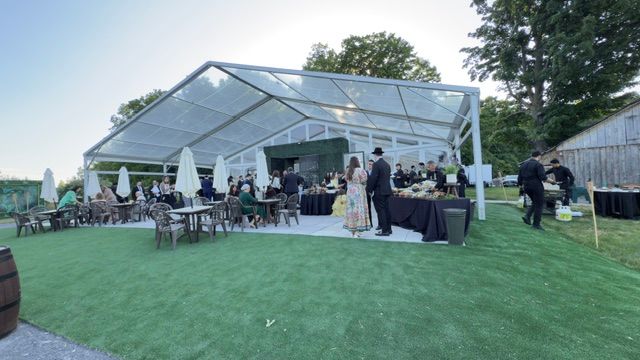 Événement en plein air sous une tente à toit transparent avec des invités ; tables avec parasols, personnes qui discutent, tables de restauration et un espace gazonné.
