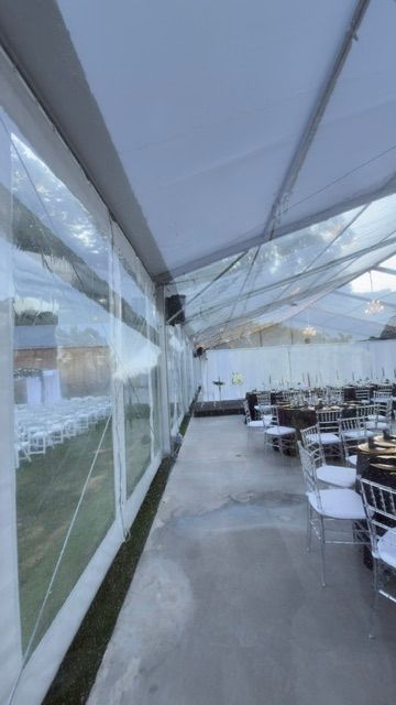 À l'intérieur d'une tente événementielle aux parois transparentes ; rangées de tables et de chaises sur un sol poli, paroi latérale donnant sur l'extérieur.