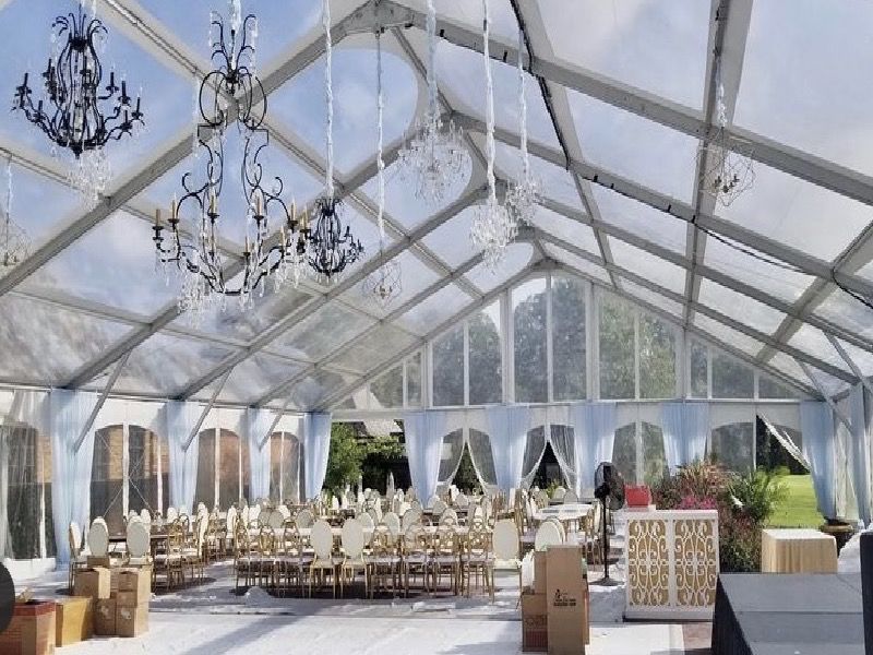 Aménagement intérieur d'une tente transparente pour un événement, avec tables, chaises, lustres et vue sur l'extérieur.