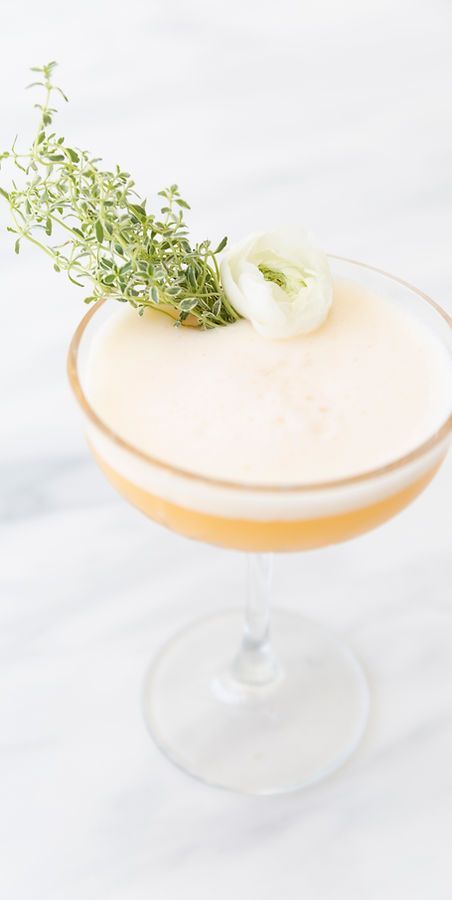 Cocktail servi dans un verre à pied, garni de thym et de fleur blanche, sur un plateau en marbre blanc.