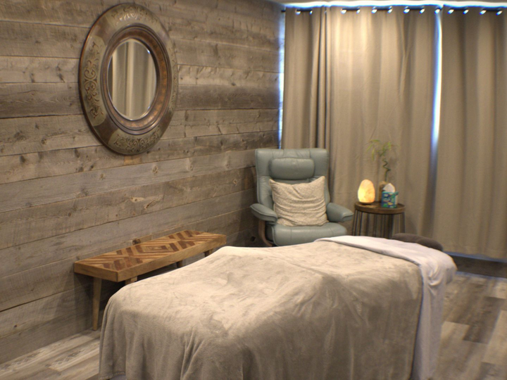 Massage room with a massage table, chair, mirror, and salt lamp. Wooden wall and beige curtains.