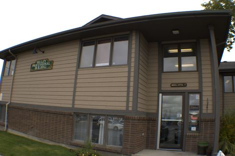 Brown building with dark trim and brick base, a sign above the door, and windows.