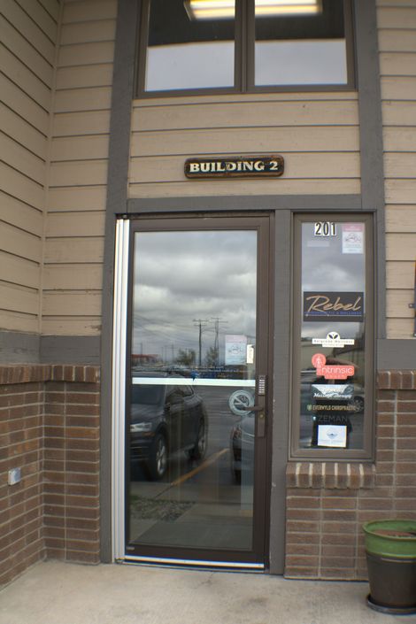 Building entrance with glass door and storefront window; brown trim, brick facade, cloudy sky reflection.