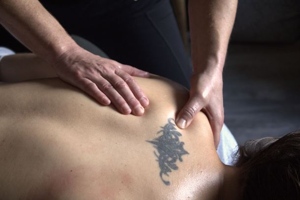 Hands giving a back massage on a person's upper back and shoulder. A tattoo is visible.