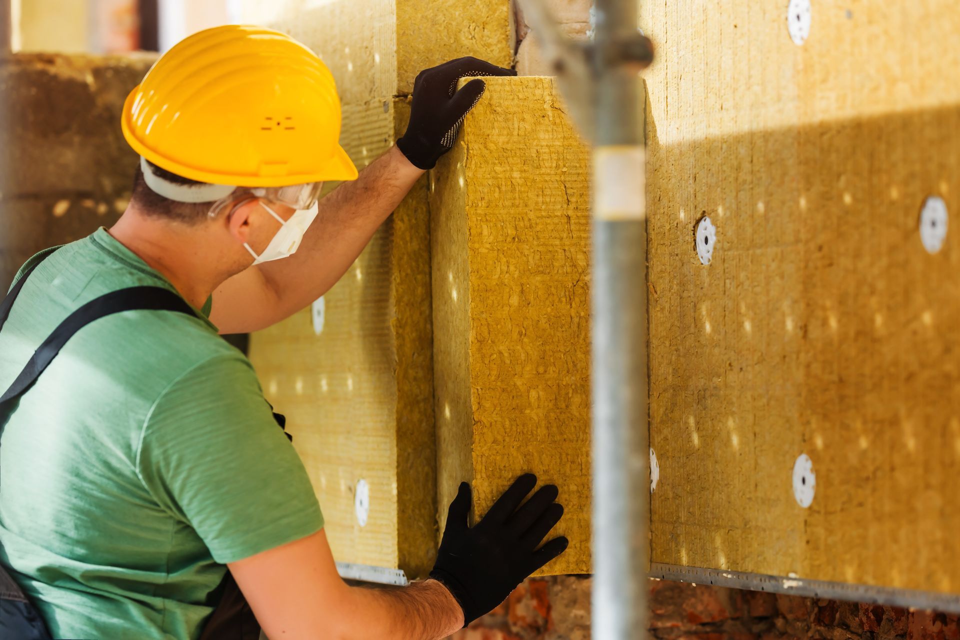 Construction worker installing insulation on a wall; wearing a hard hat, gloves, and mask.