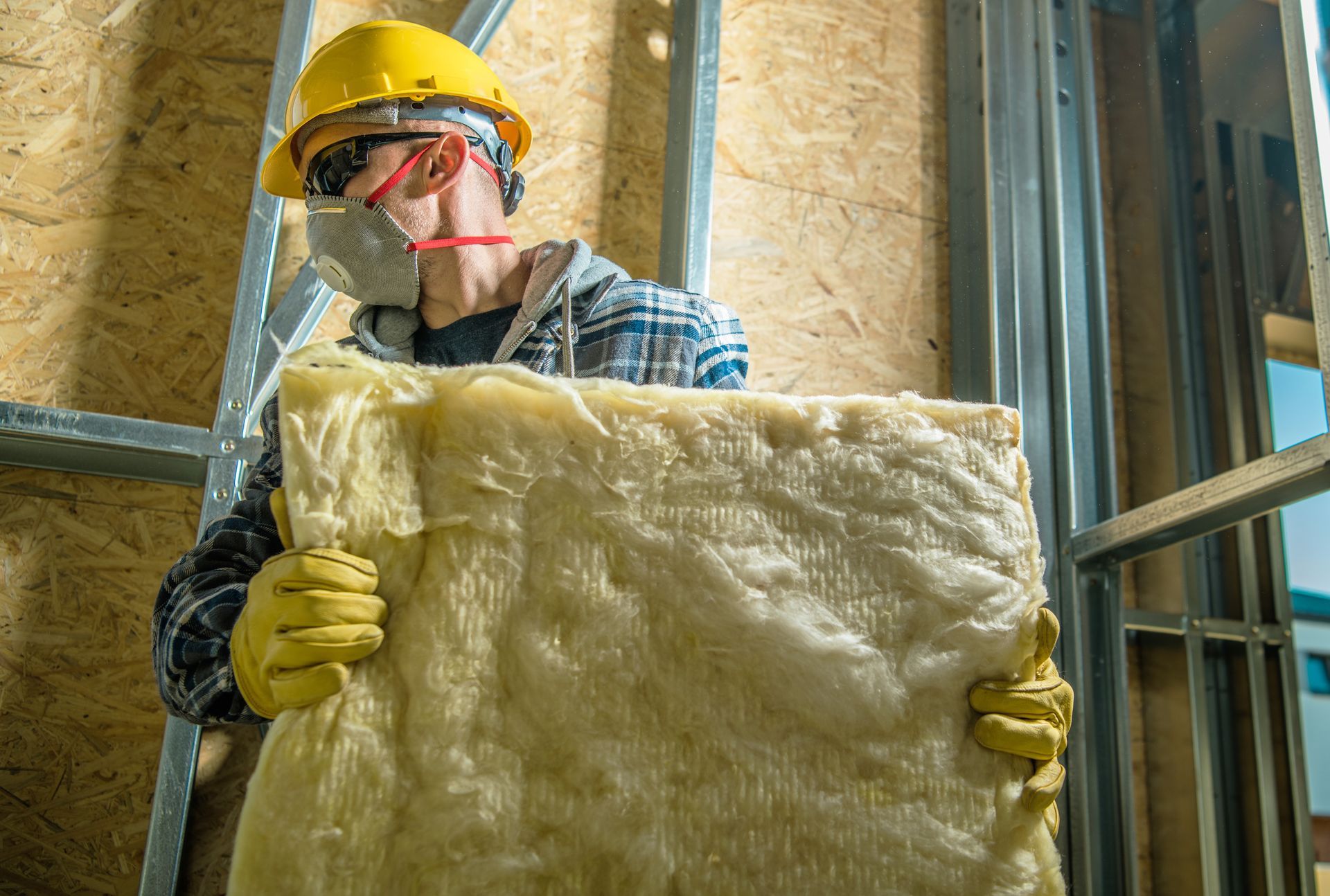 Construction worker in hard hat and gloves holding insulation batts inside a framed wall.