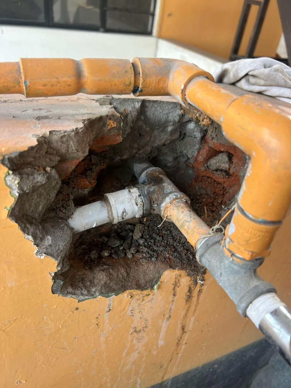 Pared dañada que deja expuestas las tuberías. Tuberías anaranjadas y blancas, ladrillos y escombros visibles.