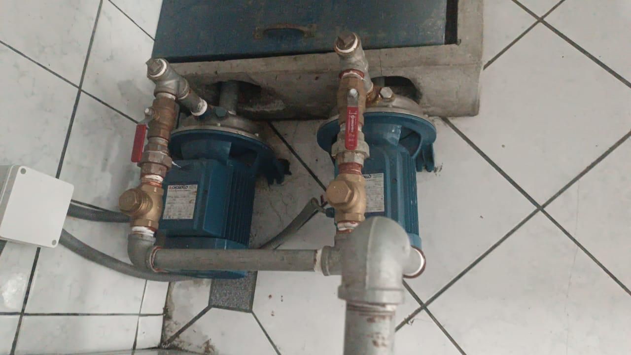 Bombas de agua azules con válvulas rojas y tuberías de metal unidas a un aparato gris montado sobre azulejos blancos.