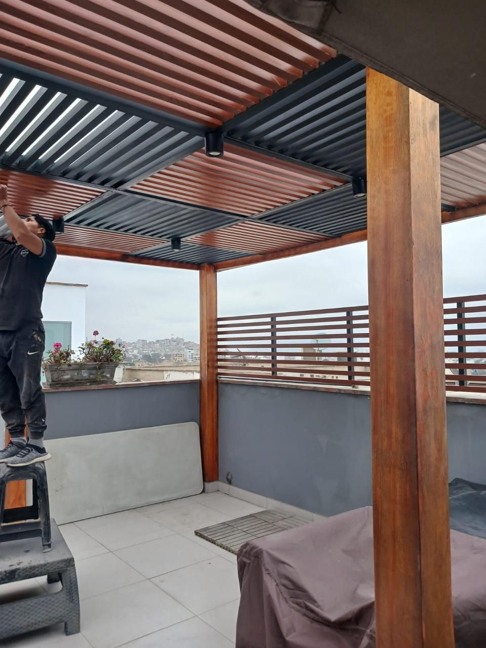 Persona instalando una pérgola en un patio. Postes de madera marrón y listones metálicos grises y marrones.