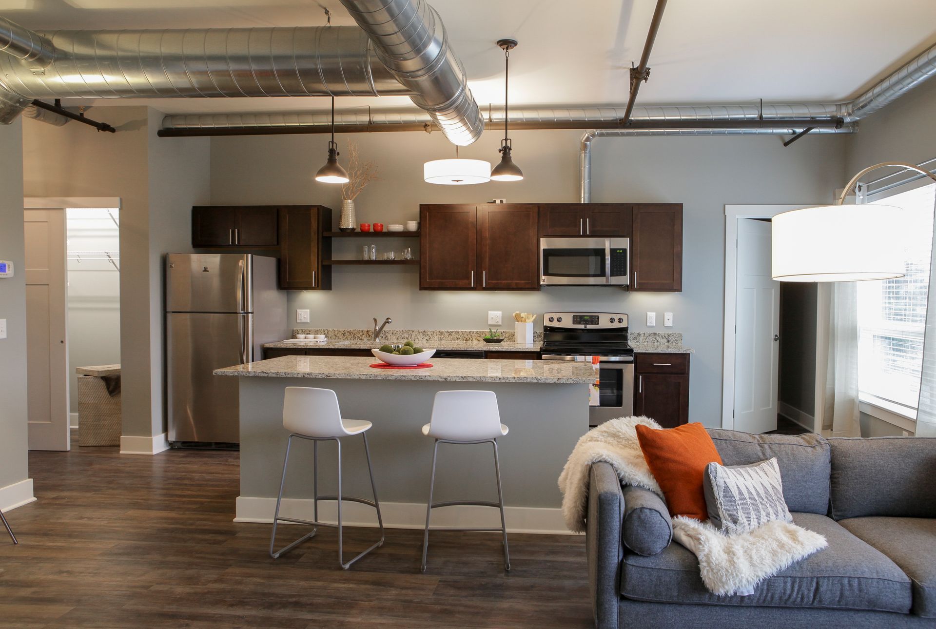 Modern open-concept kitchen and living room with stainless appliances, gray sofa, and bar stools under pendant lights