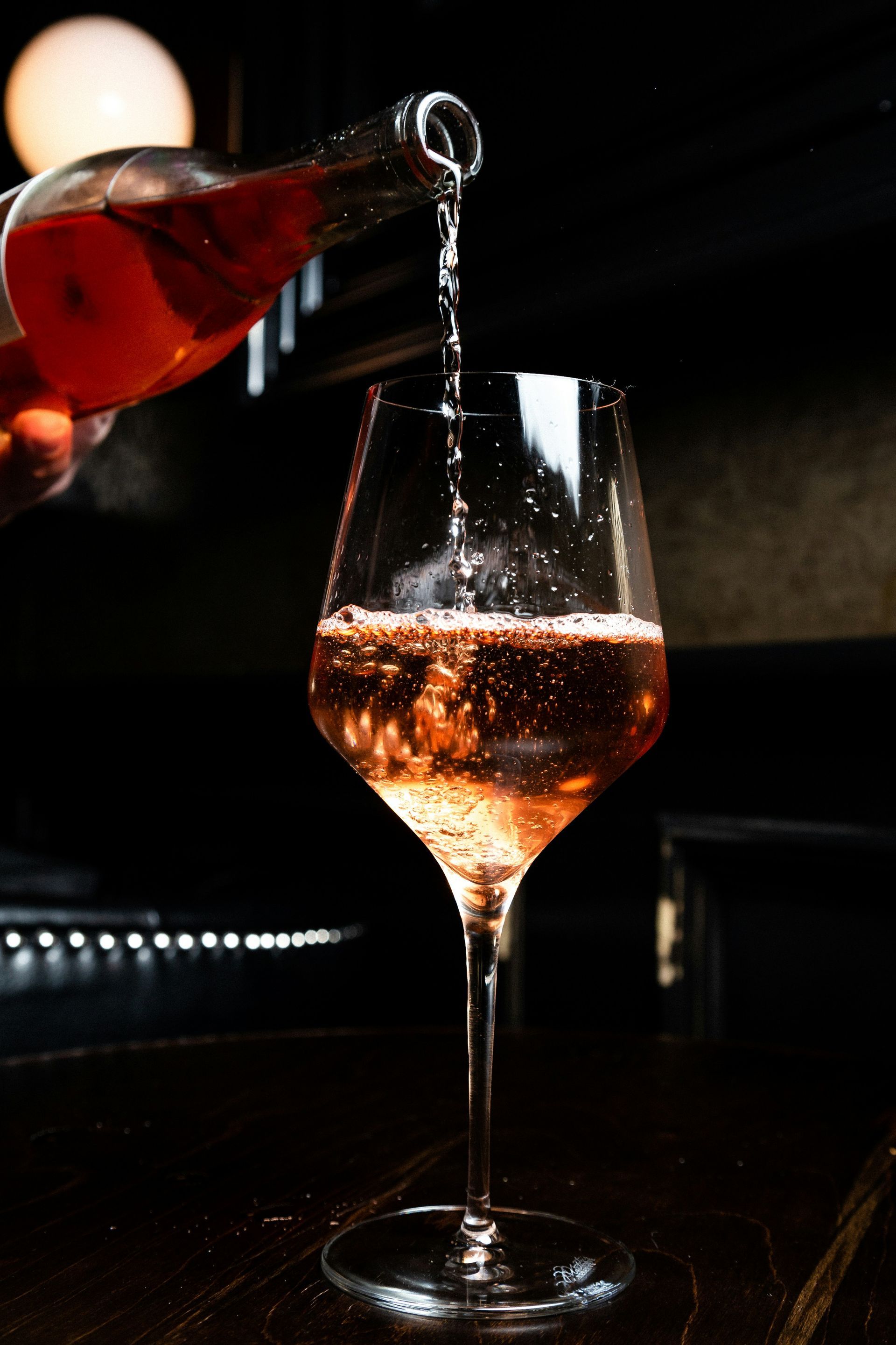 Red wine being poured into a glass on a dark bar table under dim lighting