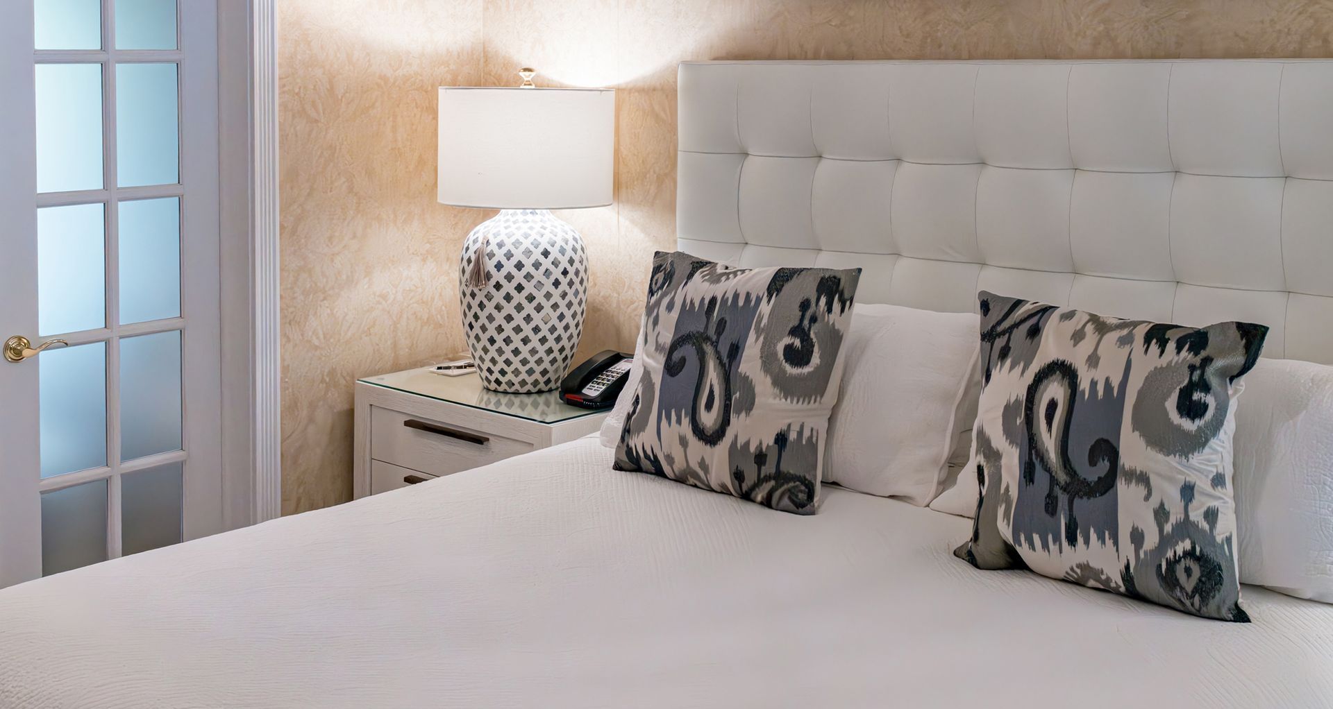 A hotel room with a bed , nightstand , lamp and door.