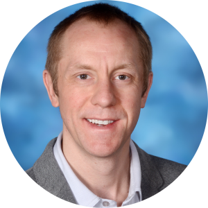 Man with fair skin, smiling, wearing a gray blazer and collared shirt, against a blue background.