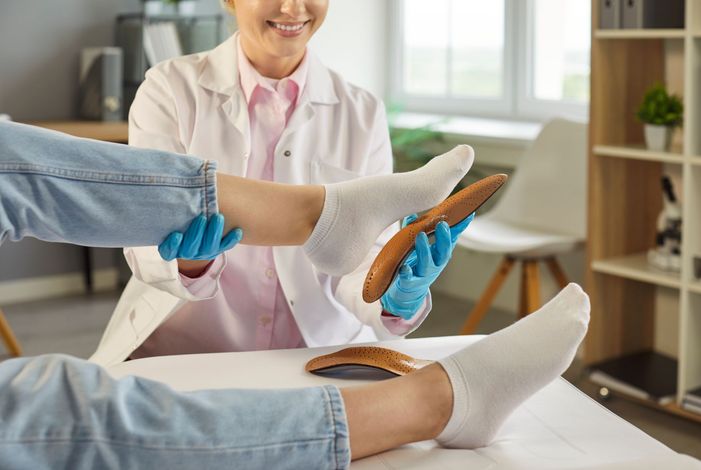 A doctor examines a patient's feet with wooden orthotics, wearing gloves and a white coat, in a clinic.