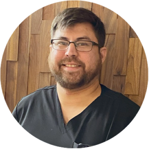 Man with glasses and beard, smiling, wearing a black shirt, in front of wood-paneled wall.