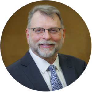 Smiling man wearing glasses, suit, and patterned tie. Oval portrait with neutral background.