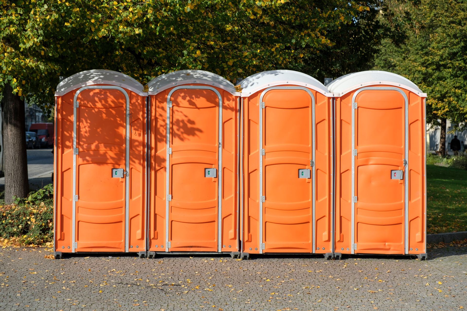 Four orange portable toilets lined up side-by-side on a gravel path near trees. Four orange portable toilets lined up side-by-side on a gravel path near trees.