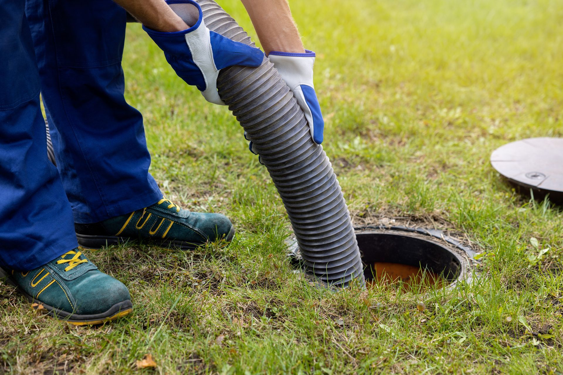 A Worker inserts a hose into an open septic tank on a grassy field.