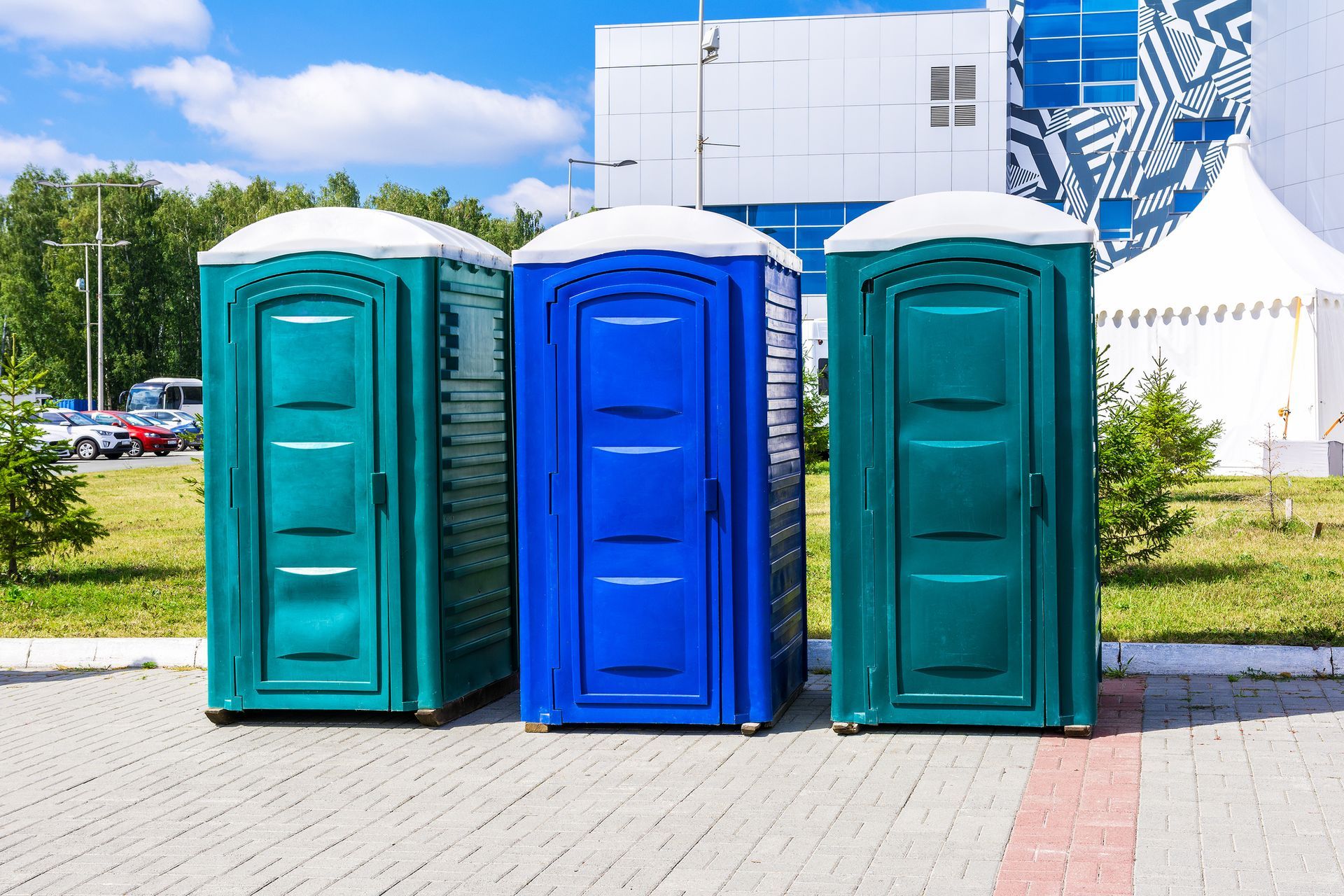 Three portable toilets are lined up on a sidewalk in front of a building.