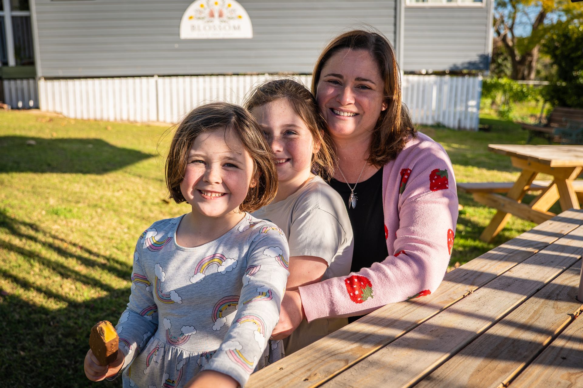 Woman and two girls smile, posing near a picnic table outside a light gray building.