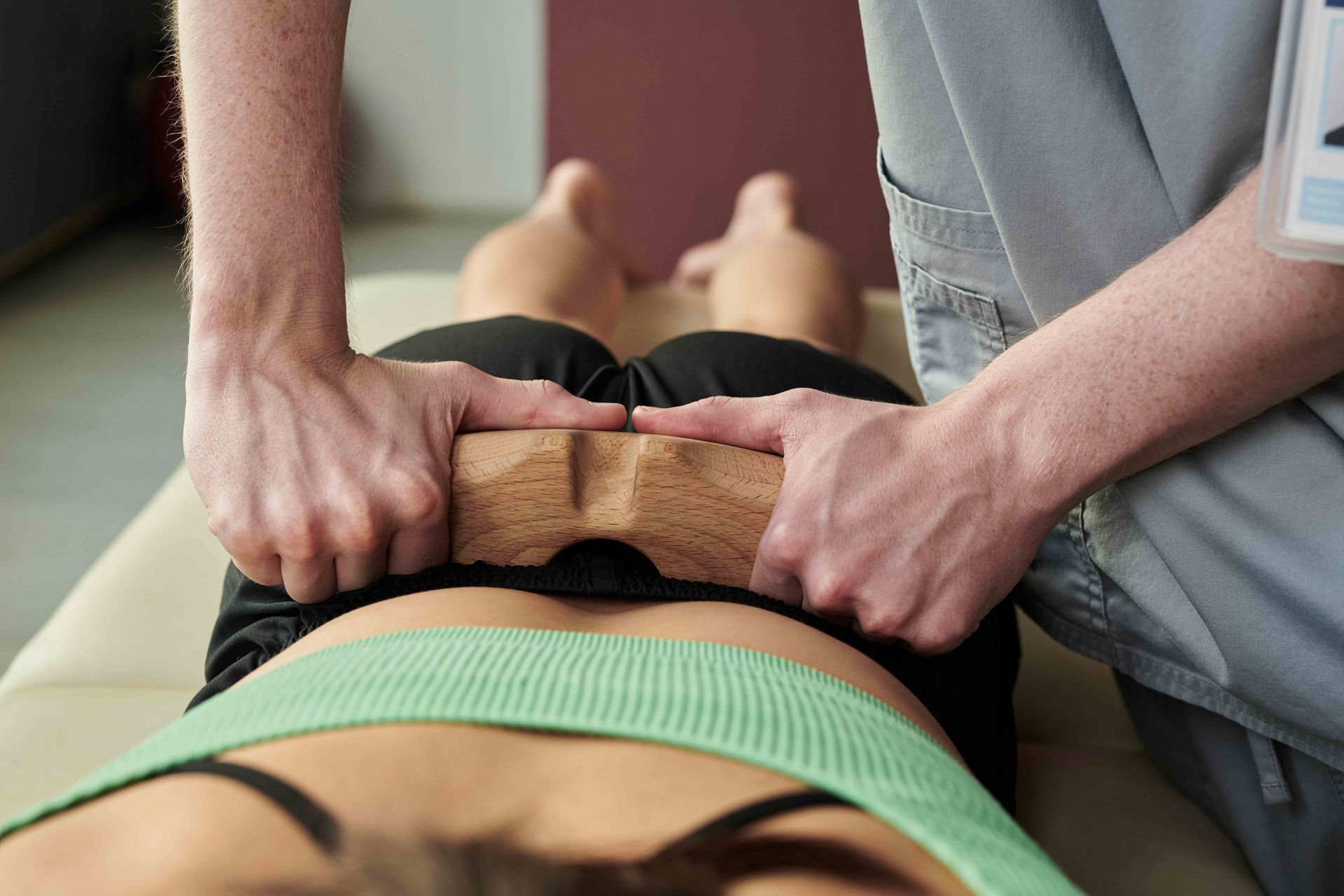 Person's back being massaged with a wooden tool on a massage table. The person's wearing a green top.