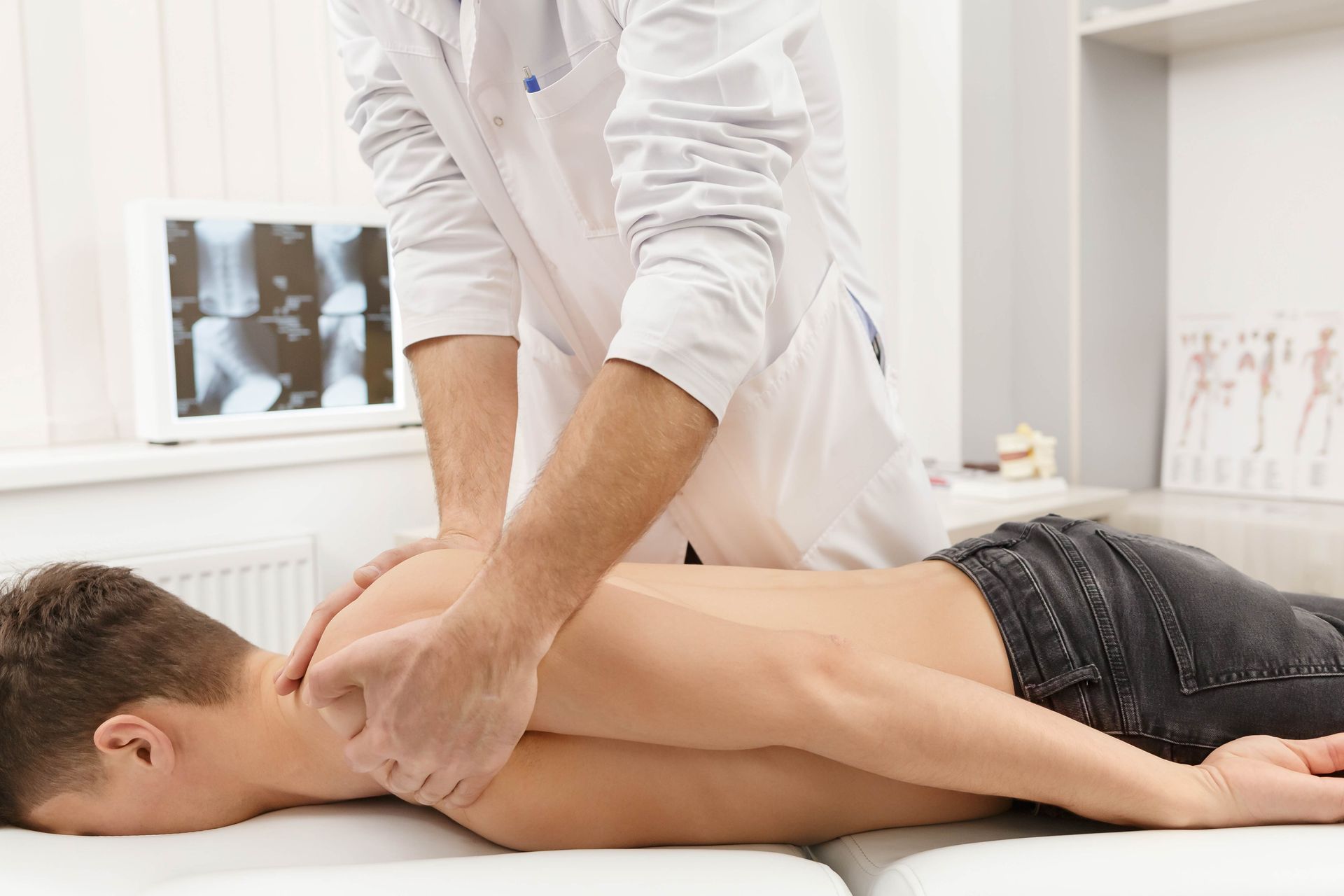 Doctor examining patient's back on a medical table, with X-ray in the background.