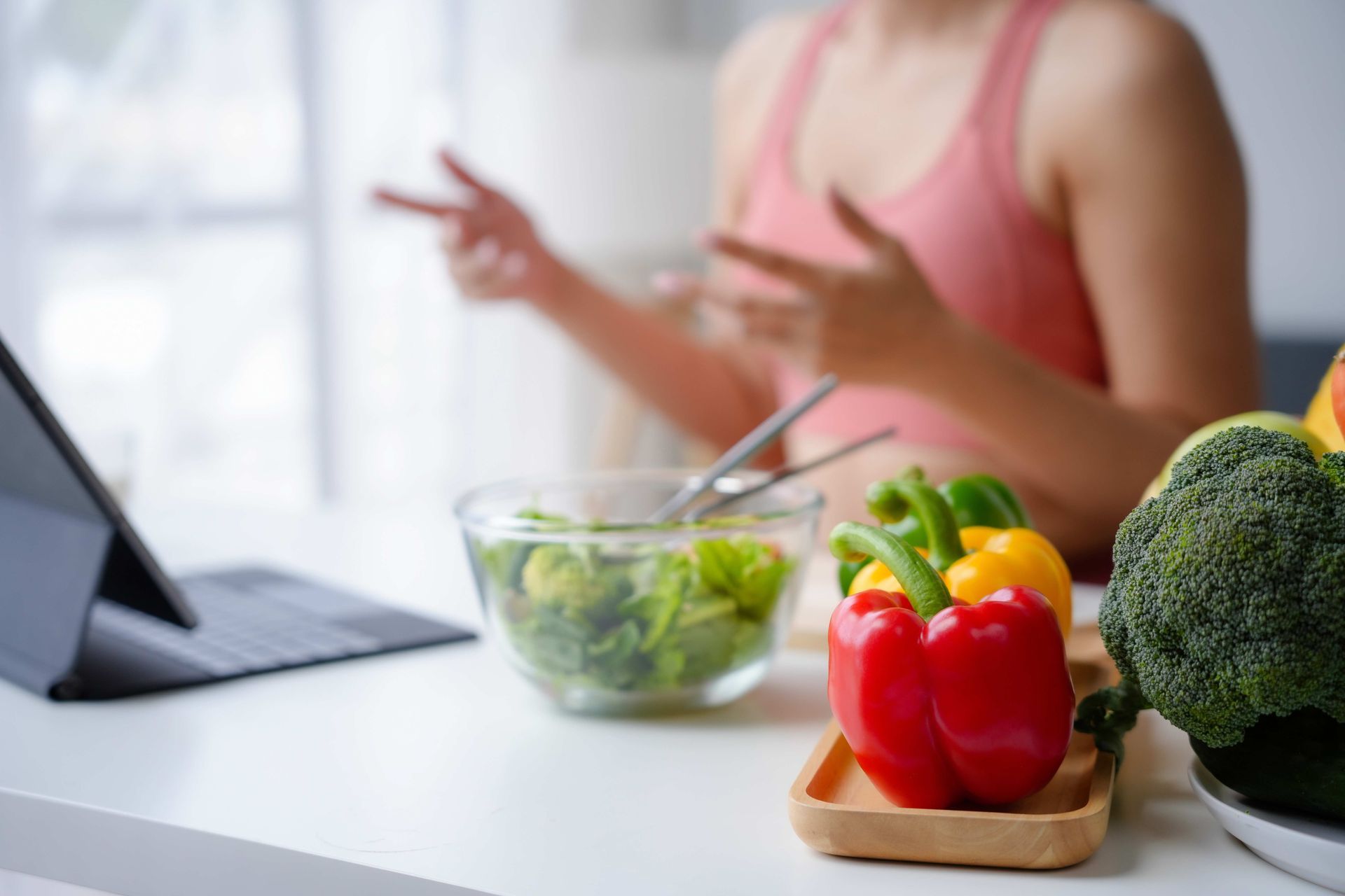 Woman in pink top gestures, a laptop, and vegetables on a white table.