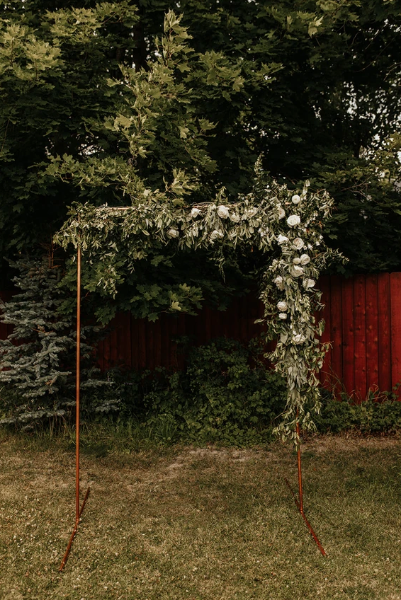 A wooden arch decorated with white flowers is sitting in the grass in front of a red fence.