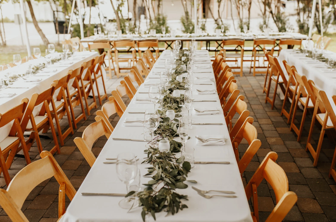 A long table with a white tablecloth and wooden chairs is set up for a wedding reception.