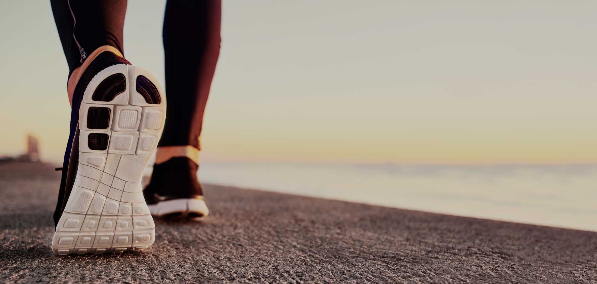 Runner's feet in sneakers on a path by the water, sunset in background.