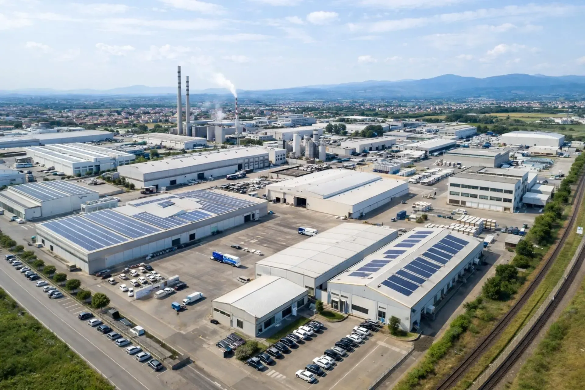 Vista aerea di un complesso industriale con numerosi edifici, alcuni dotati di pannelli solari, e ciminiere sullo sfondo.